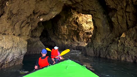 Slow-motion-pov-shot-from-boat-following-couple-in-kayak-cruising-into-cliff-cave-at-Algarve-coastline,Portugal