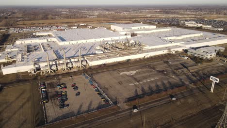 Aerial-orbital-shot-of-the-huge-Ford-Mustang-assembly-plant-in-Michigan-with-cars-passing-by-on-a-highway-and-printed-logo-on-the-car-park