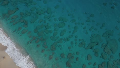 Aerial-top-down-shot-of-rocks-on-ground-of-blue-colored-Caribbean-sea-with-sandy-beach-in-sun