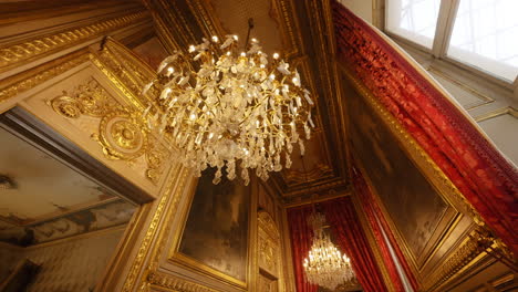 Vintage-illuminated-chandeliers-hanging-from-a-rich-ceiling-with-paintings-and-decorations,-inside-the-Louvre-Museum,-France