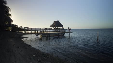 Abenddämmerung-Am-Strand-Von-Punta-Gorda,-Während-Die-Silhouette-Zweier-Kinder-Am-Rand-Des-Piers-In-Roatan,-Honduras,-Spielt-–-Handheld