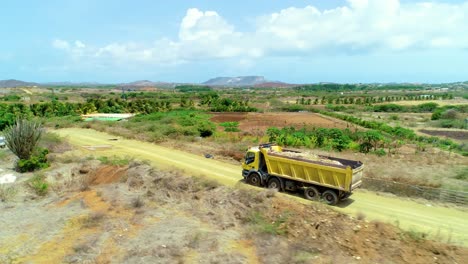 4k-aerial-cinematic-follow-orbit-shot-of-yellow-dump-truck-filled-with-dirt-and-rubbish,-driving-through-rural-farmland-on-a-dirt-road-during-a-beautiful-sunny-day-in-the-Caribbean,-Curacao