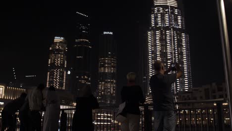 People,-tourists,-families-watching-the-water-fountain,-light-show-at-the-Dubai-mall-in-Downtown-Dubai,-UAE-with-skyscrapers-and-buildings-lit-up-at-night