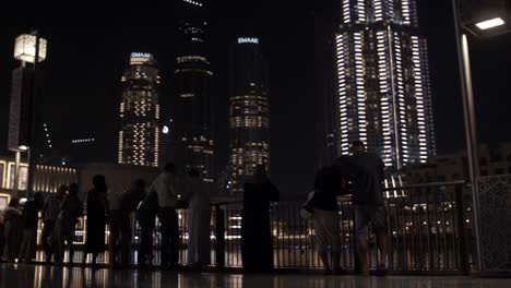 People-watching-the-water-fountain,-light-show-at-the-Dubai-mall-with-distant,-lit-up-buildings-and-skyscrapers