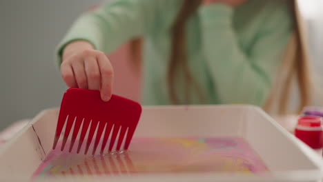 Girl-uses-comb-to-draw-on-oily-water-surface-in-tray-at-desk
