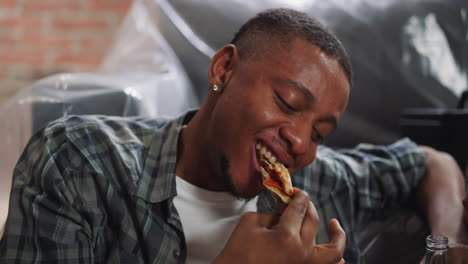 Smiling-Afro-American-guy-bites-pizza-slice-leaning-on-sofa