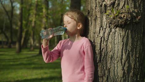 Tired-little-girl-drinks-water-leaning-on-old-tree-in-park