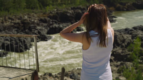 Woman-adjusts-hair-standing-on-rocky-bank-of-mountain-river