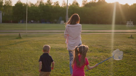 Lady-with-playful-children-walks-along-park-field-at-sunset