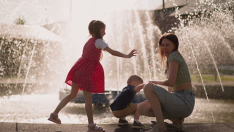 Girl-splashes-water-in-mother-squatting-with-son-by-fountain