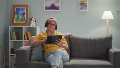 Old-lady-with-headphones-surfs-internet-via-tablet-at-home
