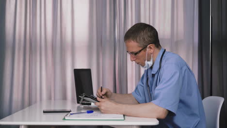 doctor-in-uniform-and-facial-mask-writes-at-table-in-office