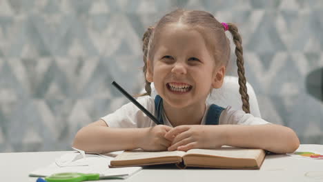 Glückliches-Kleines-Mädchen-Macht-Hausaufgaben-Mit-Buch-Am-Tisch