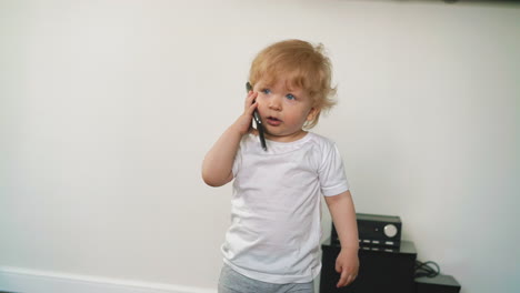 Un-Niño-Pequeño-Con-Camiseta-Habla-En-Un-Teléfono-Inteligente-Cerca-De-Una-Pared-Blanca
