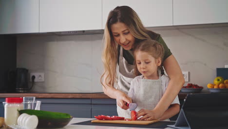 mother-teaches-little-girl-in-apron-to-cut-tomato-at-table