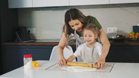 mother-and-little-daughter-roll-dough-at-table-in-kitchen