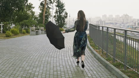 Positive-woman-with-umbrella-runs-on-city-park-at-rain