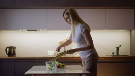 Lady-pours-cut-cucumbers-into-glass-bowl-at-table-in-kitchen
