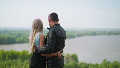 Blonde-woman-and-motorcyclist-hug-looking-at-river