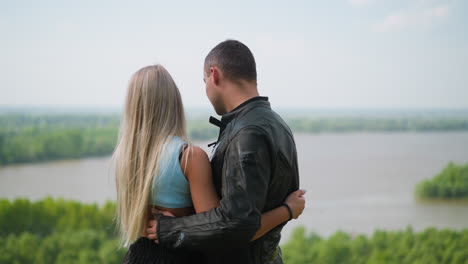 Couple-embraces-and-stands-on-hill-against-picturesque-lake