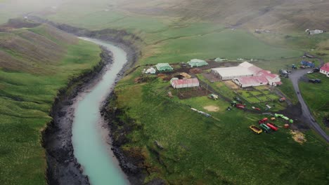 Vista-Aérea-De-Una-Hermosa-Granja-Islandesa-Encaramada-Justo-Encima-De-Un-Río-Glacial-Azul-Y-Campos-Verdes