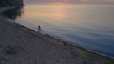 Aerial-follows-woman-walking-on-beach-solo-as-sunset-glow-reflects-on-water