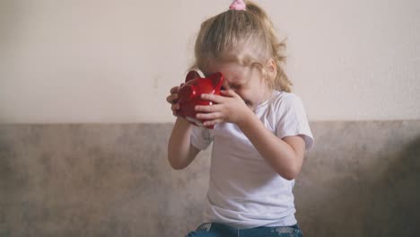 little-girl-in-white-looks-into-red-piggybank-hole-on-top