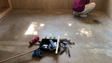 Painting-doing-home-improvement-diy-remodel-of-basement-with-light-wood-panels-and-concrete-floor-painting-bright-white-husband-and-wife-prepping-ground-and-walls---in-4K-60fps