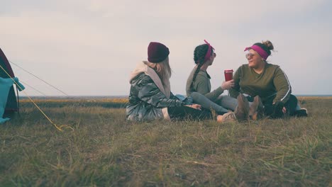 happy-women-with-drinks-look-at-sky-sitting-on-grass-by-tent