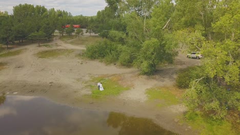 newlywed-couple-walks-along-bank-of-calm-lake-aerial-view
