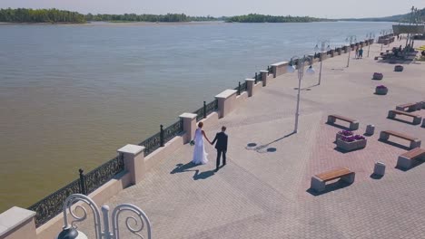 just-married-couple-walks-along-waterfront-near-river-aerial