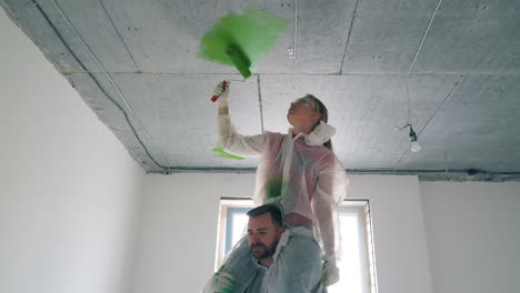 man-holds-woman-painting-ceiling-with-green-color-in-room