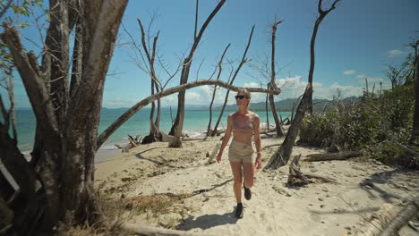 Female-traveler-explores-tropical-sand-beach-with-dead-trees-on-shore