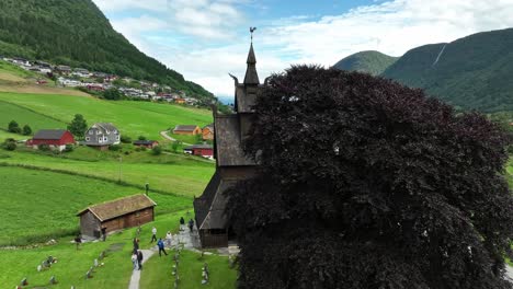 Iglesia-De-Madera-De-Hopperstad-En-Vik-Sogn-Noruega---órbita-Aérea-Que-Revela-El-Edificio-Detrás-De-Un-árbol---Turistas-En-Visita-Guiada-Caminando-Afuera---60-Fps
