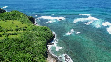 Bird's-eye-view-above-rolling-green-grassy-hill-with-tropical-trees-and-waves-crashing-over-rocks