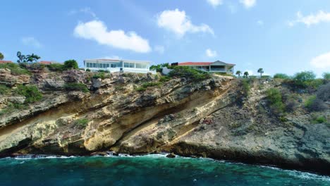 Drone-rises-towards-angled-eroded-rock-of-caribbean-island,-stunning-home-on-cliff-edge