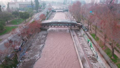 Flyover-over-the-Mapocho-River-with-a-large-amount-of-water,-Cultural-Center-of-the-Bridge-Theater