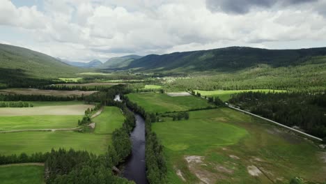 Aerial-View-Of-River-Flowing-Through-The-Green-Fields-With-Mountains-In-Daytime