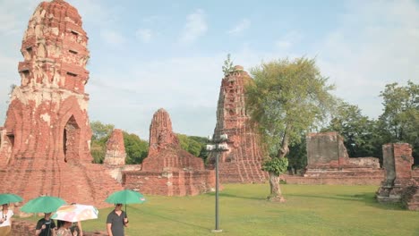 Turistas-Caminando-Por-Wat-Maha,-Las-Ruinas-Del-Templo-Tailandés-En-Ayutthaya-En-Un-Día-Caluroso-Con-Sombrillas