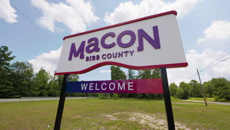 Macon-Bibb-County-Georgia-welcome-road-sign-in-front-of-highway-road-woods-wide-angle-pan-down-right-sunny-summer-sky-clouds-60p-0001
