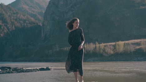graceful-woman-stands-on-rock-at-river-against-mountains