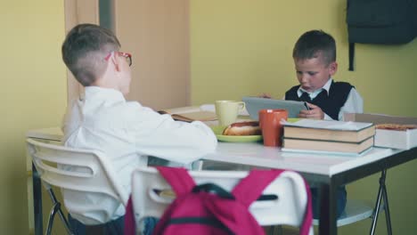 little-boy-plays-on-tablet-schoolmate-sits-at-white-table