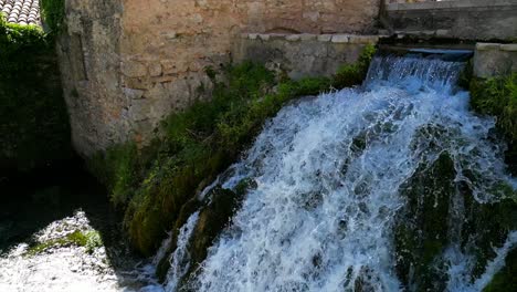 Aerial-View-Of-Waterfall-Flowing-Past-Building-Into-River-In-Rasiglia-Village-Town