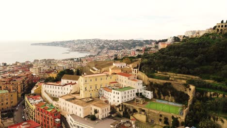 Aerial-backwards-shot-showing-soccer-field-on-top-of-hill-with-beautiful-coastline-of-Naples-at-cloudy-day---Old-town-with-historical-buildings-and-sea-in-Italy-_-establishing-drone-shot