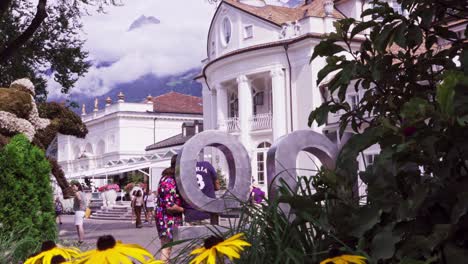 People-are-walking-along-the-spa-promenade-whilst-others-are-taking-pictures-and-selfies-in-the-city-center-of-Meran,-just-opposite-the-Kurhaus