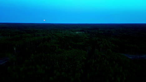 Cinematic-drone-footage-flying-over-a-forest-with-the-moon-in-the-sky-in-the-evening