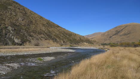 Panning-shot-of-clear-river-flowing-through-hills-and-dry-grassland