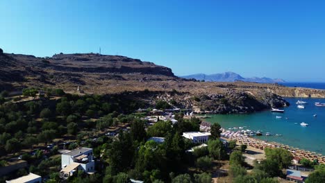 Playa-Y-Pueblo-De-Lindos-En-Rodas,-Grecia-Con-La-Acrópolis-De-Lindos,-Casas-Y-El-Mar-Mediterráneo-Durante-El-Día-Filmados-Con-El-Drone.