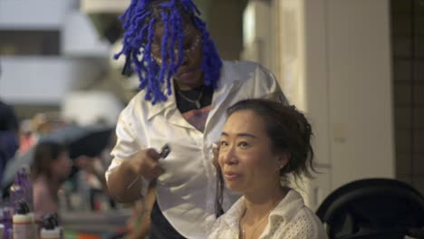 Asiatische-Frau-Lässt-Sich-Bei-Einem-Ethnischen-Kulturfestival-Im-Innenbereich-Von-Einer-Afrikanischen-Friseurin-Eine-Frische-Frisur-Machen,-Gefilmt-Als-Mittlere-Nahaufnahme-In-Zeitlupe-Im-Handheld-Stil