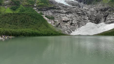 Forward-moving-aerial-close-to-green-glacial-lake-before-slowly-tilting-up-revealing-Boyabreen-glacier-Fjaerland---Part-of-Jostedal-glacier-in-Norway---Tourists-standing-in-the-edge-of-left-frame